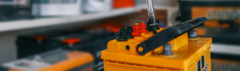A person using a hydrometer to test a yellow car battery, with visible red and black terminals. Equipment for checking battery fluid density
